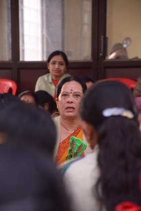 Several women are seated indoors, with one woman in the foreground wearing traditional clothing, appearing to hold a pair of glasses. The focus is on her as others are slightly blurred, possibly indicating a gathering or meeting.