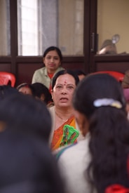 Several women are seated indoors, with one woman in the foreground wearing traditional clothing, appearing to hold a pair of glasses. The focus is on her as others are slightly blurred, possibly indicating a gathering or meeting.