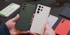 A pair of hands holding two smartphones with multiple cameras on the back, surrounded by other smartphones placed on a table. The phones are sleek, with one being dark green and the other a light cream color.