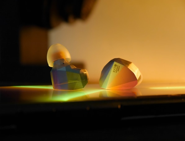 Two modern, geometric-shaped earphones are positioned on a reflective surface. The lighting casts colorful reflections and gradients across their surfaces, giving them a futuristic and artistic appearance. One earphone displays the word 'KATO' along with additional text, suggesting branding or model information.