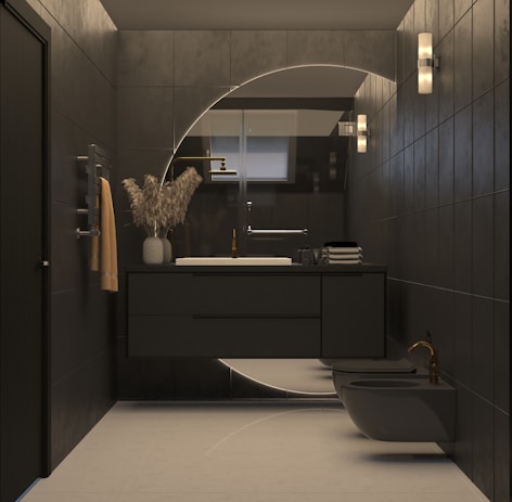 A modern bathroom with dark tiles and minimalistic design. The vanity area features a circular mirror with backlighting, a sleek black countertop with a white sink, and a brass faucet. A vase with decorative dried plants is placed on the counter alongside folded towels. Wall sconces provide additional lighting, and a neatly hung towel adds a touch of warmth to the space. The room also includes a bidet and toilet, both in dark tones.