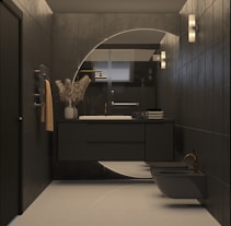 A modern bathroom with dark tiles and minimalistic design. The vanity area features a circular mirror with backlighting, a sleek black countertop with a white sink, and a brass faucet. A vase with decorative dried plants is placed on the counter alongside folded towels. Wall sconces provide additional lighting, and a neatly hung towel adds a touch of warmth to the space. The room also includes a bidet and toilet, both in dark tones.