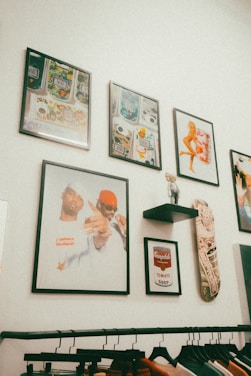 A collection of framed artwork is displayed on a white wall. The artworks include abstract patterns, a classic Campbell's Tomato Soup design, and a portrait of two individuals. There's also a skateboard mounted next to a small shelf with a toy-like figurine. Below, a clothing rack holds items on hangers.