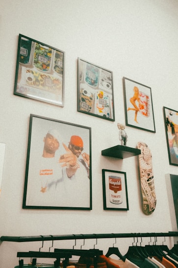 A collection of framed artwork is displayed on a white wall. The artworks include abstract patterns, a classic Campbell's Tomato Soup design, and a portrait of two individuals. There's also a skateboard mounted next to a small shelf with a toy-like figurine. Below, a clothing rack holds items on hangers.