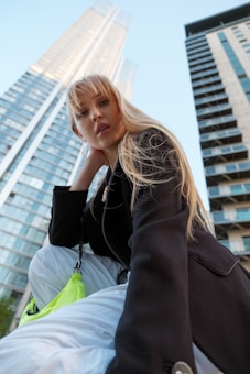 A candid photograph of a person with long blonde hair looking into the camera, set against the backdrop of tall modern skyscrapers. The person is wearing a black jacket and holding a neon green bag, with a confident expression and relaxed posture.