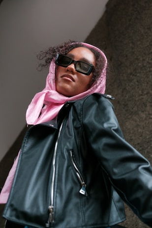 A person is wearing a pink headscarf and black leather jacket, looking up while wearing rectangular black sunglasses. The angle of the photo is from below, creating a sense of empowerment.