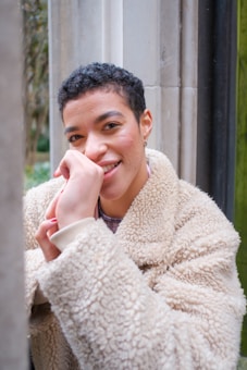 The image depicts a person with short curly hair smiling warmly at the camera. They are wearing a beige, fluffy, fleece jacket. The person rests their chin on their hand, standing near a stone wall with greenery visible in the background.