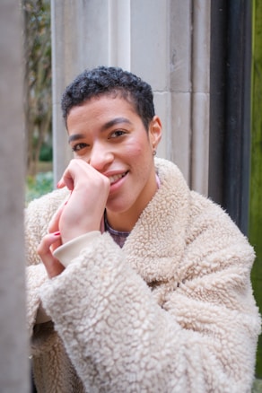 The image depicts a person with short curly hair smiling warmly at the camera. They are wearing a beige, fluffy, fleece jacket. The person rests their chin on their hand, standing near a stone wall with greenery visible in the background.