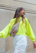 A person with long, wavy hair is looking to the side while wearing a bright neon yellow oversized sweater and white pants. They have a beige crossbody bag with two colorful circular patches, one red and one blue. The background features a textured, vertical stone wall.