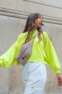 A person with long, wavy hair is looking to the side while wearing a bright neon yellow oversized sweater and white pants. They have a beige crossbody bag with two colorful circular patches, one red and one blue. The background features a textured, vertical stone wall.