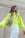 A person with long, wavy hair is looking to the side while wearing a bright neon yellow oversized sweater and white pants. They have a beige crossbody bag with two colorful circular patches, one red and one blue. The background features a textured, vertical stone wall.