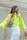 A person with long, wavy hair is looking to the side while wearing a bright neon yellow oversized sweater and white pants. They have a beige crossbody bag with two colorful circular patches, one red and one blue. The background features a textured, vertical stone wall.