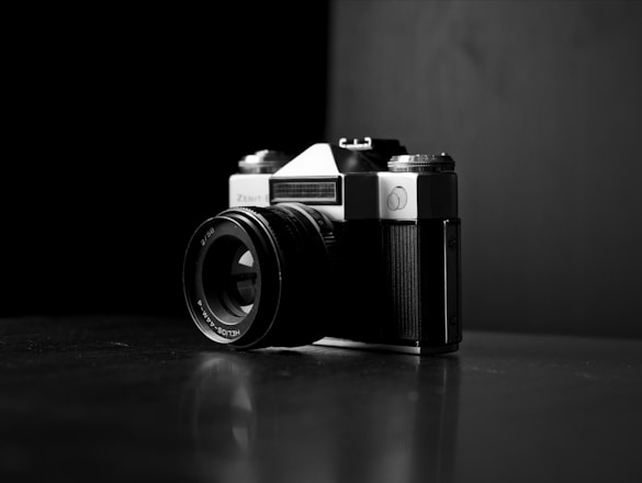 A vintage camera with a lens and visible brand markings, positioned at an angle on a reflective dark surface. The backdrop is stark and minimal, enhancing the focus on the camera itself. The interplay of light and shadow creates a dramatic and classic atmosphere.
