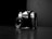 A vintage camera with a lens and visible brand markings, positioned at an angle on a reflective dark surface. The backdrop is stark and minimal, enhancing the focus on the camera itself. The interplay of light and shadow creates a dramatic and classic atmosphere.
