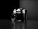 A vintage camera with a lens and visible brand markings, positioned at an angle on a reflective dark surface. The backdrop is stark and minimal, enhancing the focus on the camera itself. The interplay of light and shadow creates a dramatic and classic atmosphere.