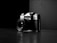 A vintage camera with a lens and visible brand markings, positioned at an angle on a reflective dark surface. The backdrop is stark and minimal, enhancing the focus on the camera itself. The interplay of light and shadow creates a dramatic and classic atmosphere.