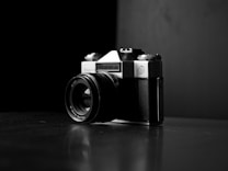 A vintage camera with a lens and visible brand markings, positioned at an angle on a reflective dark surface. The backdrop is stark and minimal, enhancing the focus on the camera itself. The interplay of light and shadow creates a dramatic and classic atmosphere.