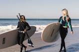 two women in wetsuits carrying surfboards on the beach