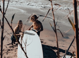 A man and woman are situated on a sandy beach near the ocean, preparing a surfboard. The woman is crouching and facing the camera, while the man, shirtless, is adjusting the large white board labeled 'Cruiser.' The scene is framed by reeds in the foreground, providing a natural filter.