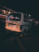 A white van is parked on a street at night, under the glow of streetlights. The background features light streaks from passing vehicles and illuminated signs, indicating a busy urban environment.