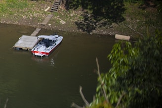 A small boat with a red and white exterior is moored next to a simple wooden dock on a calm river. The surrounding area is lush with greenery, including trees and bushes along the riverbank. A wooden staircase leads down to the dock from the land.