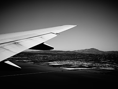 A black and white photograph capturing the wing of an airplane in flight. The wing extends across the frame with a landscape visible below, showing a mix of fields, water, and distant mountains under a clear sky.