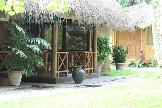 A small, thatched roof hut made from wood and bamboo with a balcony. Lush potted plants and greenery surround the structure, which sits on a stone path. A clay pot is placed on the ground, and the setting is tranquil and natural.