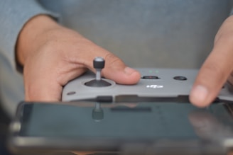 A close-up of hands holding a gray drone controller with one hand operating a joystick and the other hand touching an adjacent button. A reflective screen is visible at the bottom, possibly a smartphone display.