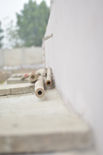 Several cylindrical pipes are laid on a rough concrete surface near a white wall. The background is blurred, showing indistinct greenery and some structures.