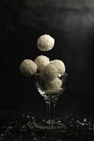 A glass cup is filled with round coconut-covered sweets, with several more appearing to hover above it. The background is dark, highlighting the white texture of the confectioneries, and small coconut flakes are scattered around the base.