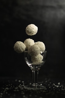 A glass cup is filled with round coconut-covered sweets, with several more appearing to hover above it. The background is dark, highlighting the white texture of the confectioneries, and small coconut flakes are scattered around the base.
