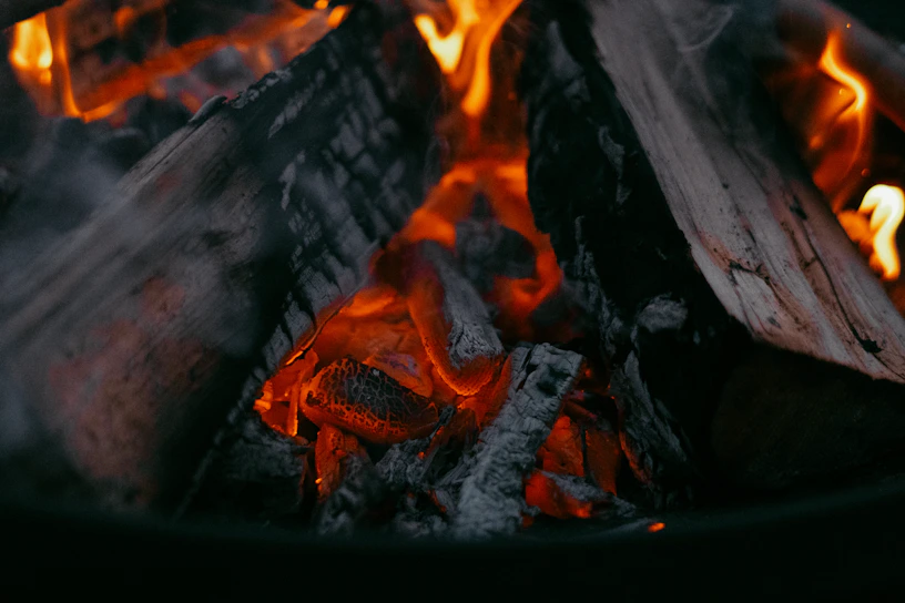 A close-up of glowing embers in a rustic fire pit, casting warm light on charred wood.