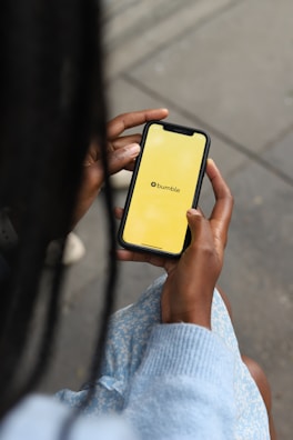 A person is holding a smartphone displaying a yellow screen with the Bumble logo. The person is wearing a light blue, patterned dress and possibly a sweater, and the setting appears to be on a street or sidewalk.