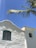 A whitewashed church with a cross on its roof against a clear blue sky. The building features an arched wooden window with a teal frame and a bell visible in a small tower. A palm tree branch extends across the top corner of the image.