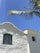 A whitewashed church with a cross on its roof against a clear blue sky. The building features an arched wooden window with a teal frame and a bell visible in a small tower. A palm tree branch extends across the top corner of the image.