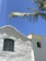 A whitewashed church with a cross on its roof against a clear blue sky. The building features an arched wooden window with a teal frame and a bell visible in a small tower. A palm tree branch extends across the top corner of the image.
