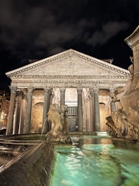 A nighttime view of an ancient Roman building featuring a grand portico with large Corinthian columns. The facade displays the historic inscription 'MAGRIPPALFCOSTERTIVMFECIT.' In front of the building, there is a fountain with ornate sculptures, including figures of sea creatures, and the water reflects a turquoise glow.