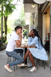 A couple sits at an outdoor café table, engaging in a lively conversation with relaxed body language. The man is wearing a white t-shirt and jeans, while the woman is in a light blue outfit. Both are smiling and leaning toward each other, surrounded by urban greenery.