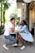 A couple sits at an outdoor café table, engaging in a lively conversation with relaxed body language. The man is wearing a white t-shirt and jeans, while the woman is in a light blue outfit. Both are smiling and leaning toward each other, surrounded by urban greenery.