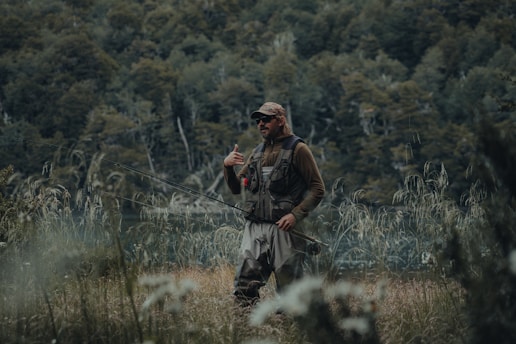 A person dressed in outdoor gear stands in a grassy area holding a fishing rod. The background features dense greenery and a body of water visible through the trees. The person appears to be in a natural and serene environment, possibly engaged in fishing.