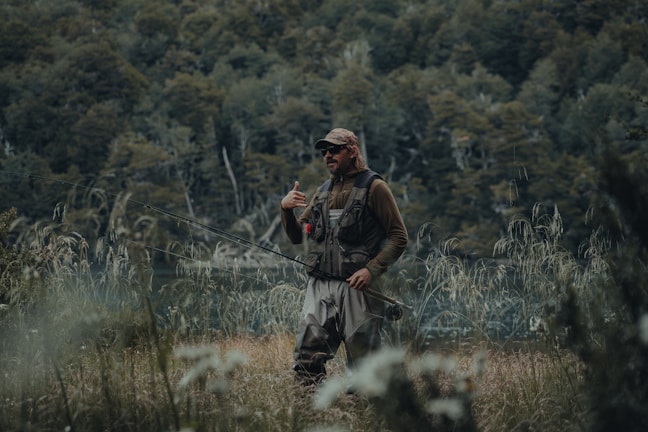 A person dressed in outdoor gear stands in a grassy area holding a fishing rod. The background features dense greenery and a body of water visible through the trees. The person appears to be in a natural and serene environment, possibly engaged in fishing.