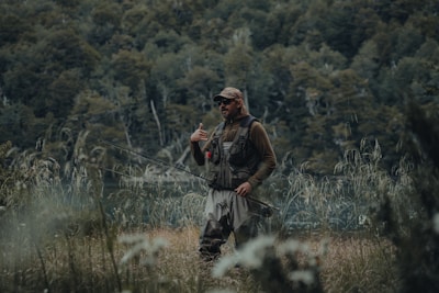 A person dressed in outdoor gear stands in a grassy area holding a fishing rod. The background features dense greenery and a body of water visible through the trees. The person appears to be in a natural and serene environment, possibly engaged in fishing.