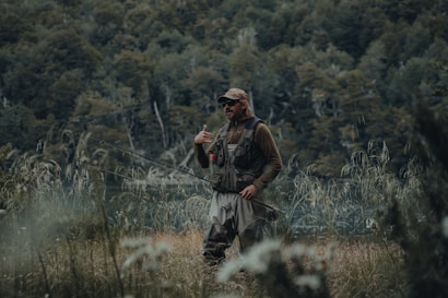 A person dressed in outdoor gear stands in a grassy area holding a fishing rod. The background features dense greenery and a body of water visible through the trees. The person appears to be in a natural and serene environment, possibly engaged in fishing.