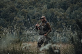 A person dressed in outdoor gear stands in a grassy area holding a fishing rod. The background features dense greenery and a body of water visible through the trees. The person appears to be in a natural and serene environment, possibly engaged in fishing.