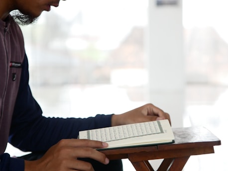 A person is sitting at a wooden stand, reading a book which appears to be a religious or academic text. The environment is bright and minimalistic, suggesting a serene or focused setting.