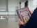 A person holds a book titled 'Syarah Rinci Rukun Iman' with a purple cover and gold lettering. The background is an indoor space with bright lighting and a polished floor, possibly a minimalist room or hall.