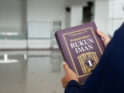 A person holds a book titled 'Syarah Rinci Rukun Iman' with a purple cover and gold lettering. The background is an indoor space with bright lighting and a polished floor, possibly a minimalist room or hall.