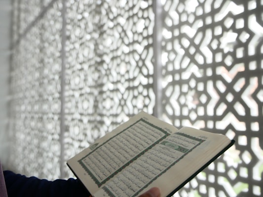 An open book with Arabic script is held by a person's hand. The background features an ornate, geometric patterned screen that casts shadows.