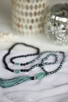 A beaded necklace with turquoise and black beads is elegantly arranged on a soft white fabric. The necklace features a prominent tassel and a larger turquoise bead. In the background, there are decorative items, including a metallic candle holder and a textured white ornament.