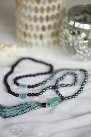 A beaded necklace with turquoise and black beads is elegantly arranged on a soft white fabric. The necklace features a prominent tassel and a larger turquoise bead. In the background, there are decorative items, including a metallic candle holder and a textured white ornament.